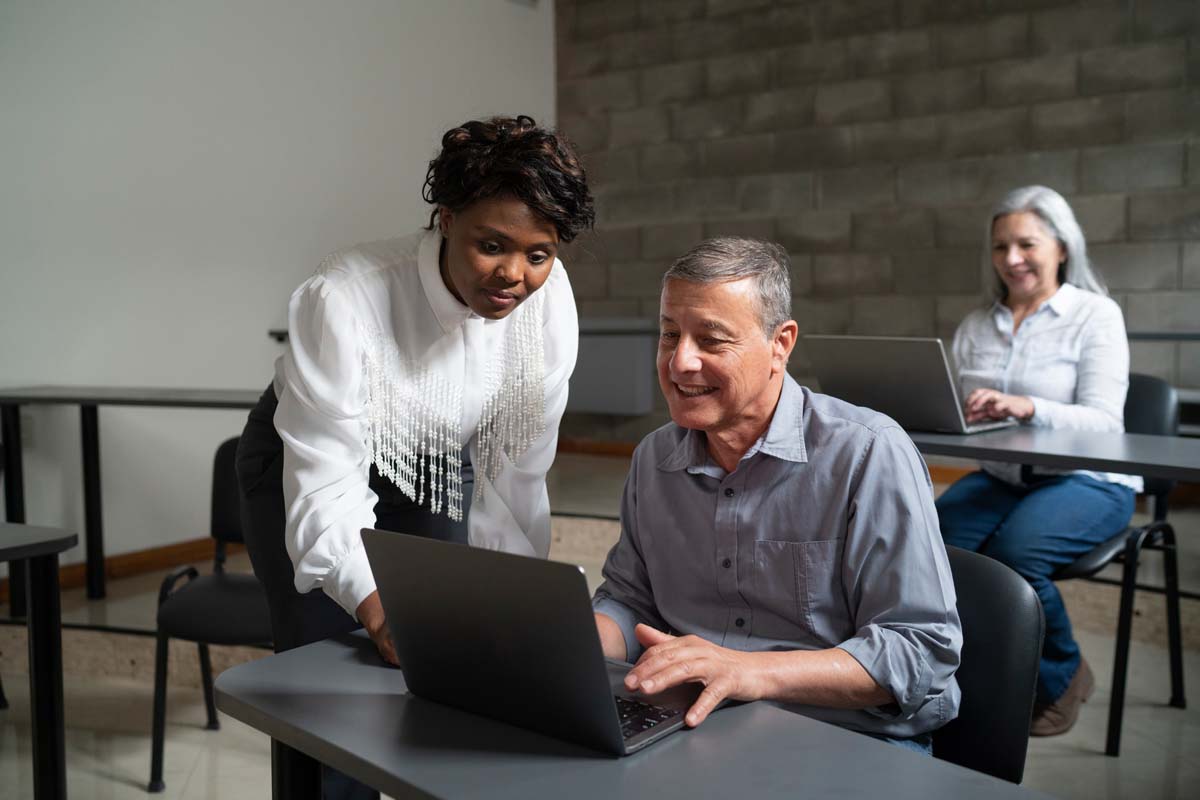 A young female African-American computing teacher is showing a student how to solve a problem on his laptop in a technology course for seniors.