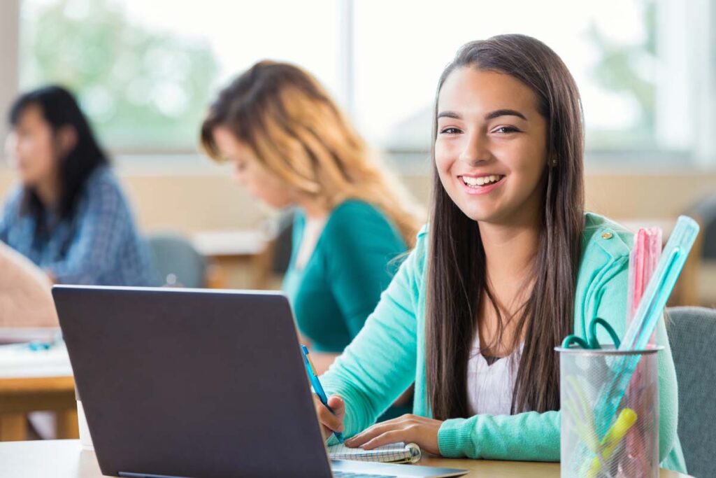 Hispanic high school student works on project in graphic design class. She is looking at the camera and smilng as she takes notes. A laptop is open on the table and art supplies are also on the table. Students are working in the background.