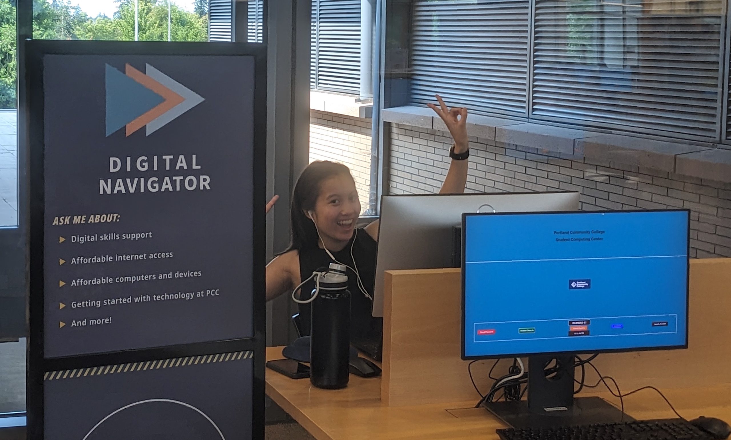 A PCC student smiles from behind a computer kiosk in a hallway next to a sign that reads Digital Navigator.