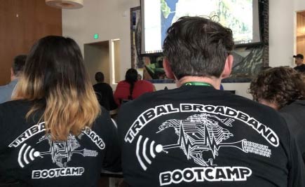 Two people watch a slideshow presentation in a large room. We see the backs of their t-shirts that read Tribal Broadband Bootcamp.