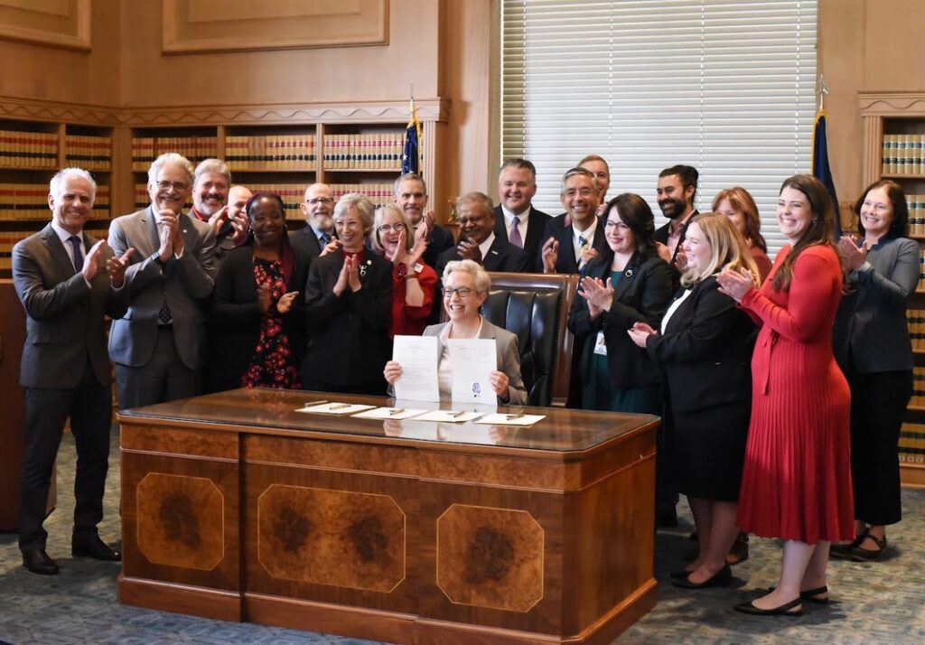 Oregon Governor Tina Kotek sits at a large wooden desk with many people standing and clapping behind her. She holds the bill she just signed into law.